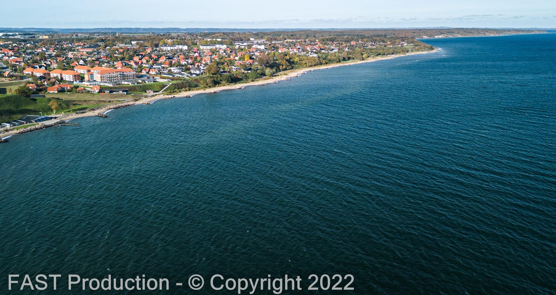 Dronefoto af kystlinje med boligkvarter og blåt hav en klar efterårsdag