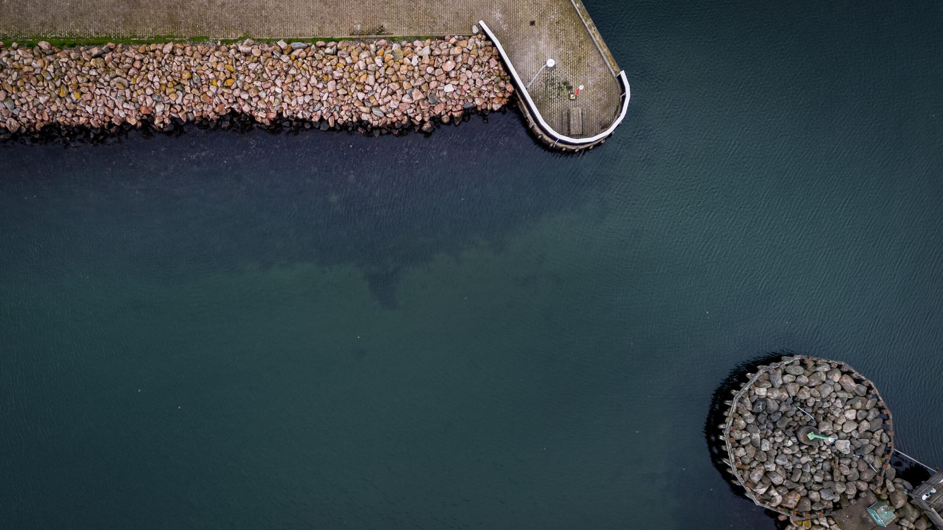 Minimalistisk dronefoto af en stensat mole og en rund stenø i dybgrønt havnevand