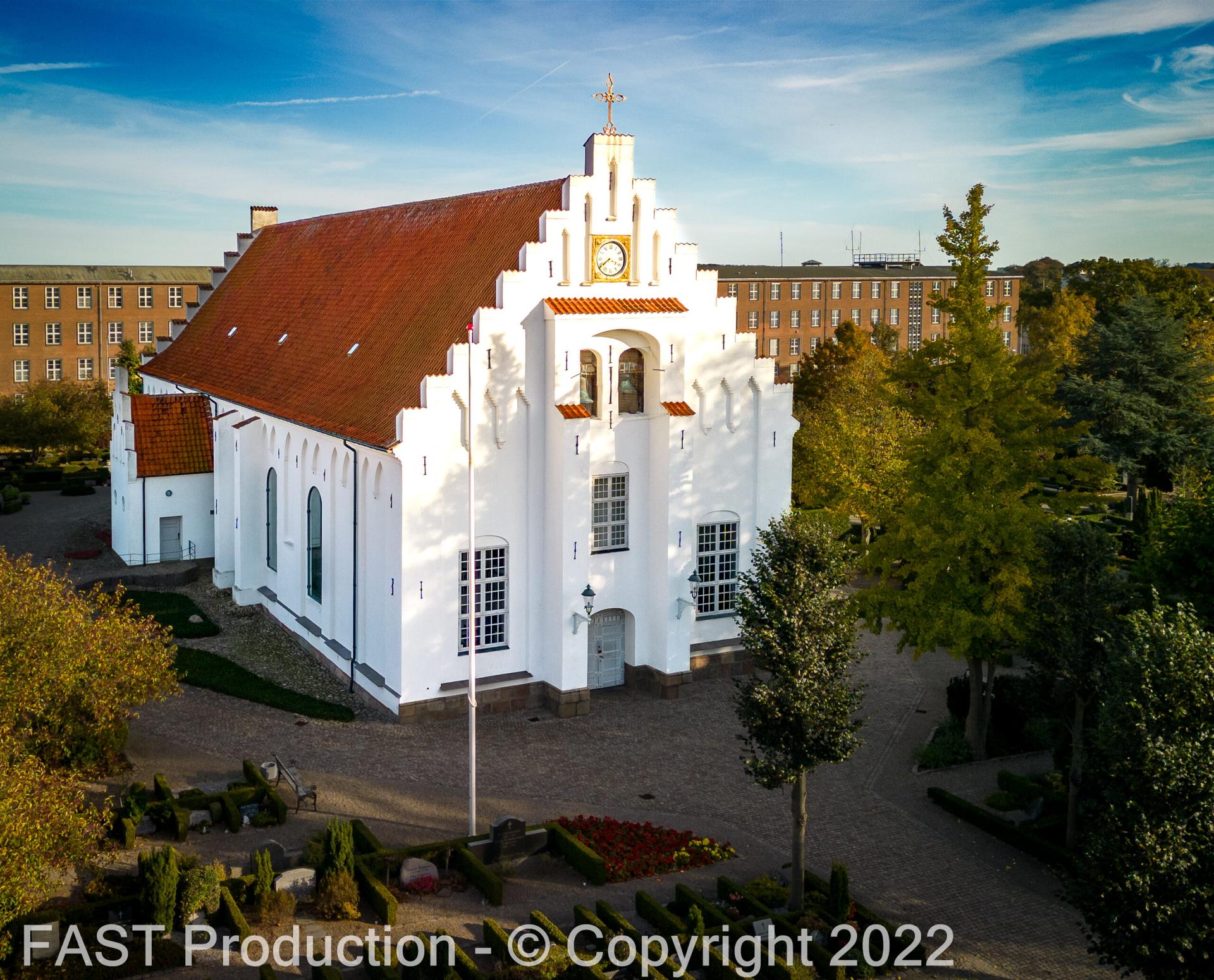 Dronefoto af hvid kirke med rødt tegltag og kamtakket gavl i efterårslys